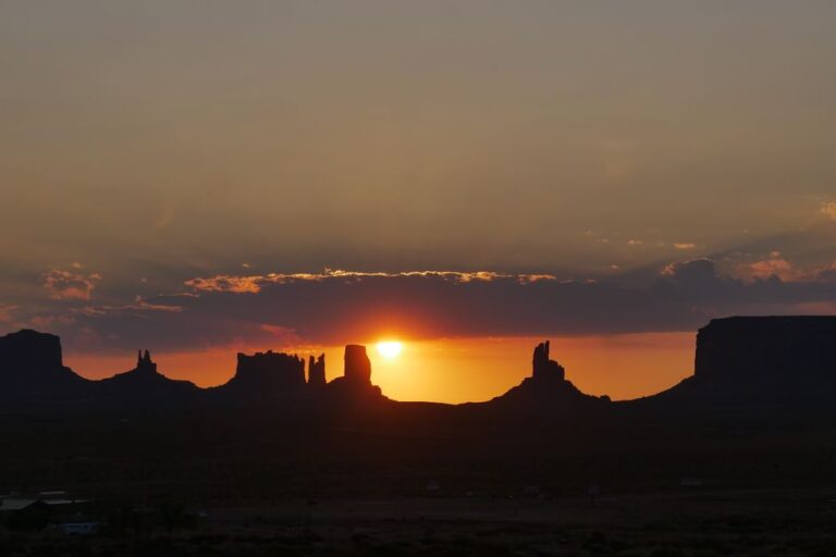 Silhouette of Monument Valley at sunset with vivid orange sky and sun peeking through rock formations.