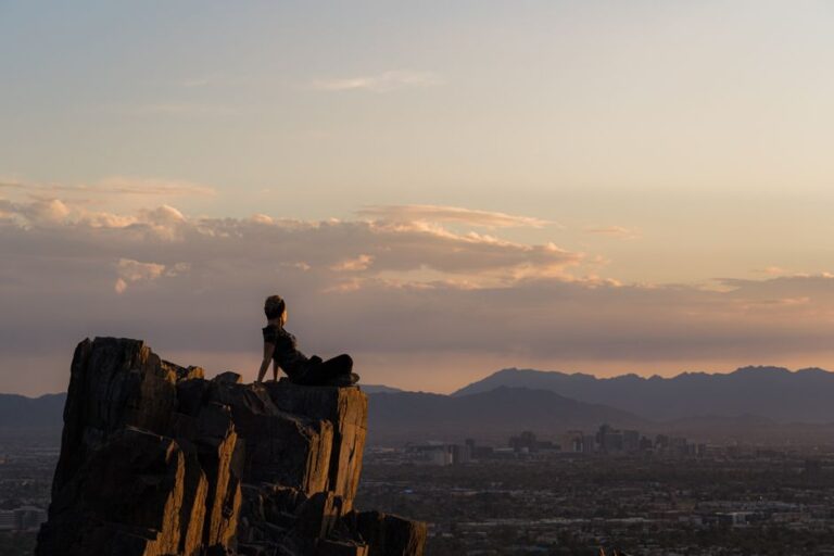 A woman sits on a rock formation overlooking Phoenix, Arizona at sunset, embracing nature and tranquility.